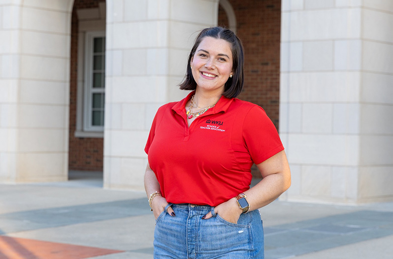 A smiling WKU student, a candidate in the alternative teacher certification program, stands outside a campus building with large columns. She is wearing a red WKU School of Teacher Education polo shirt and jeans.