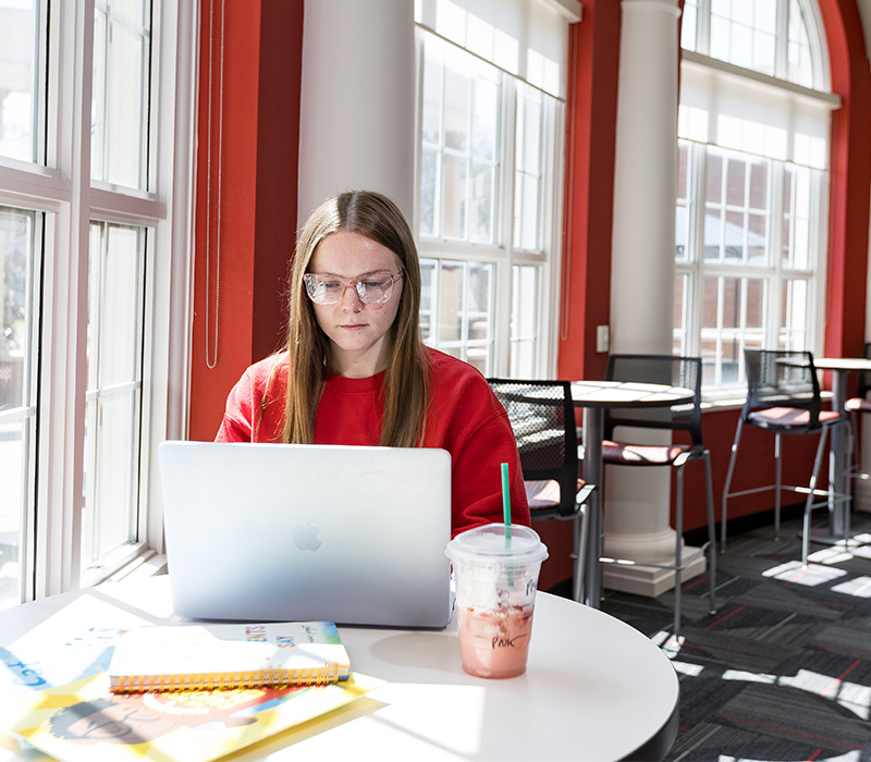 A WKU student in a red sweatshirt studying on a laptop in the sunlit CEBS Student Success Center located in Gary A. Ransdell Hall.