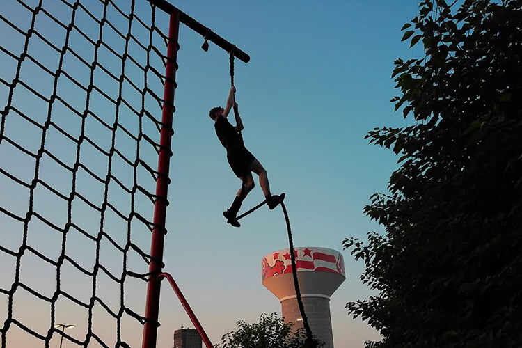 ROTC cadet climbing a vertical rope on an outdoor obstacle course during morning physical training.