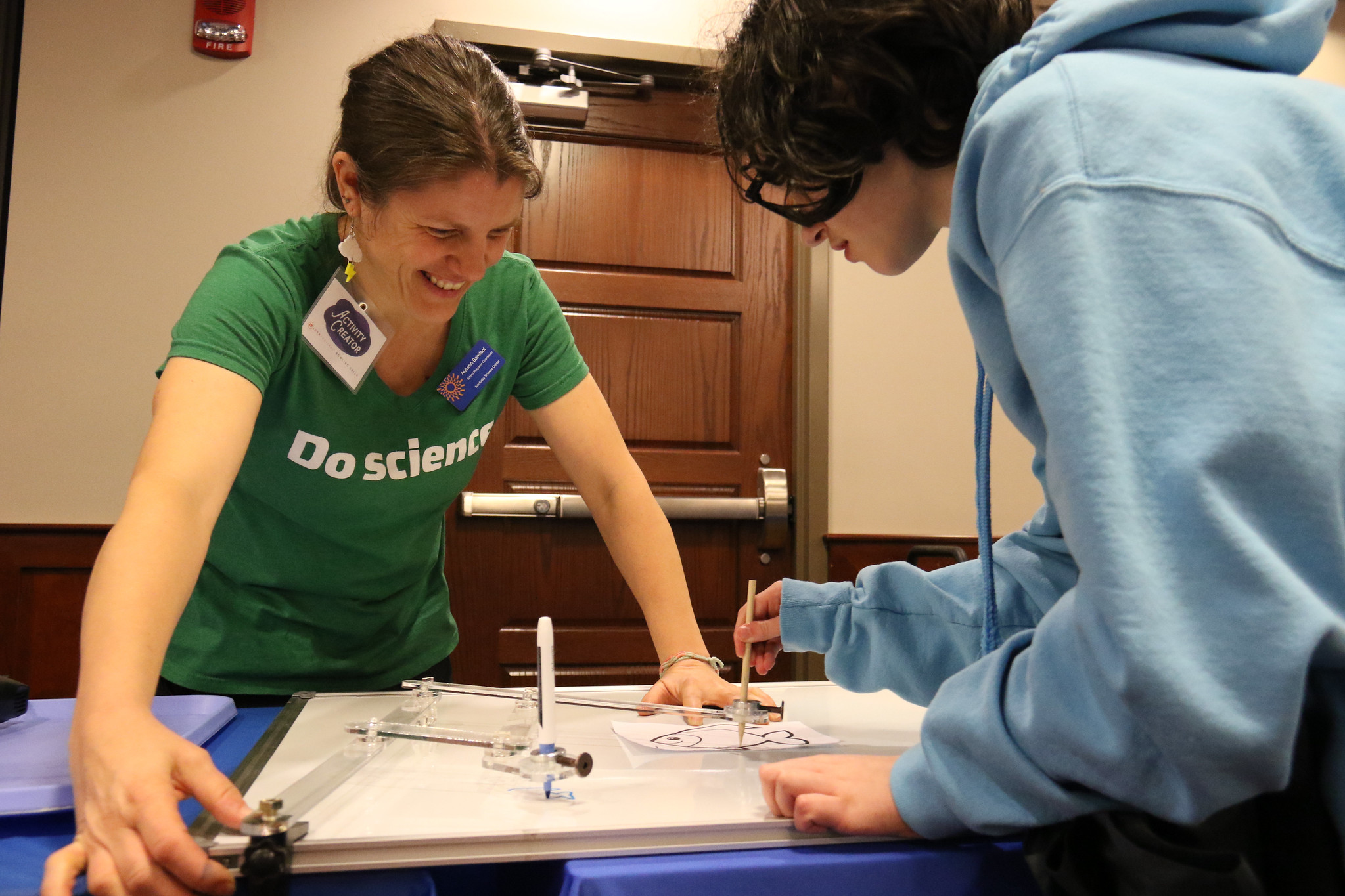 woman with arms braced on table smiling with young student wearing a blue hoodie draws on table