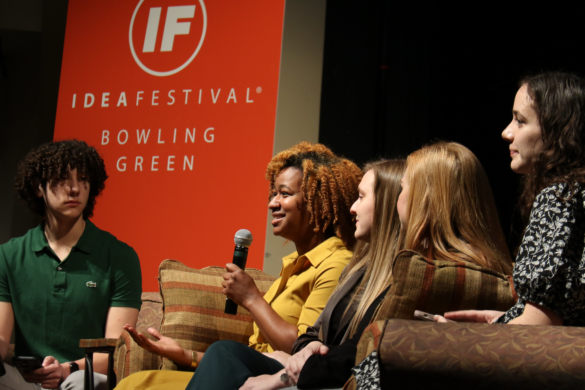 group of people sitting on couch facing to the left of the photo with orange IdeaFestival BG in background. 