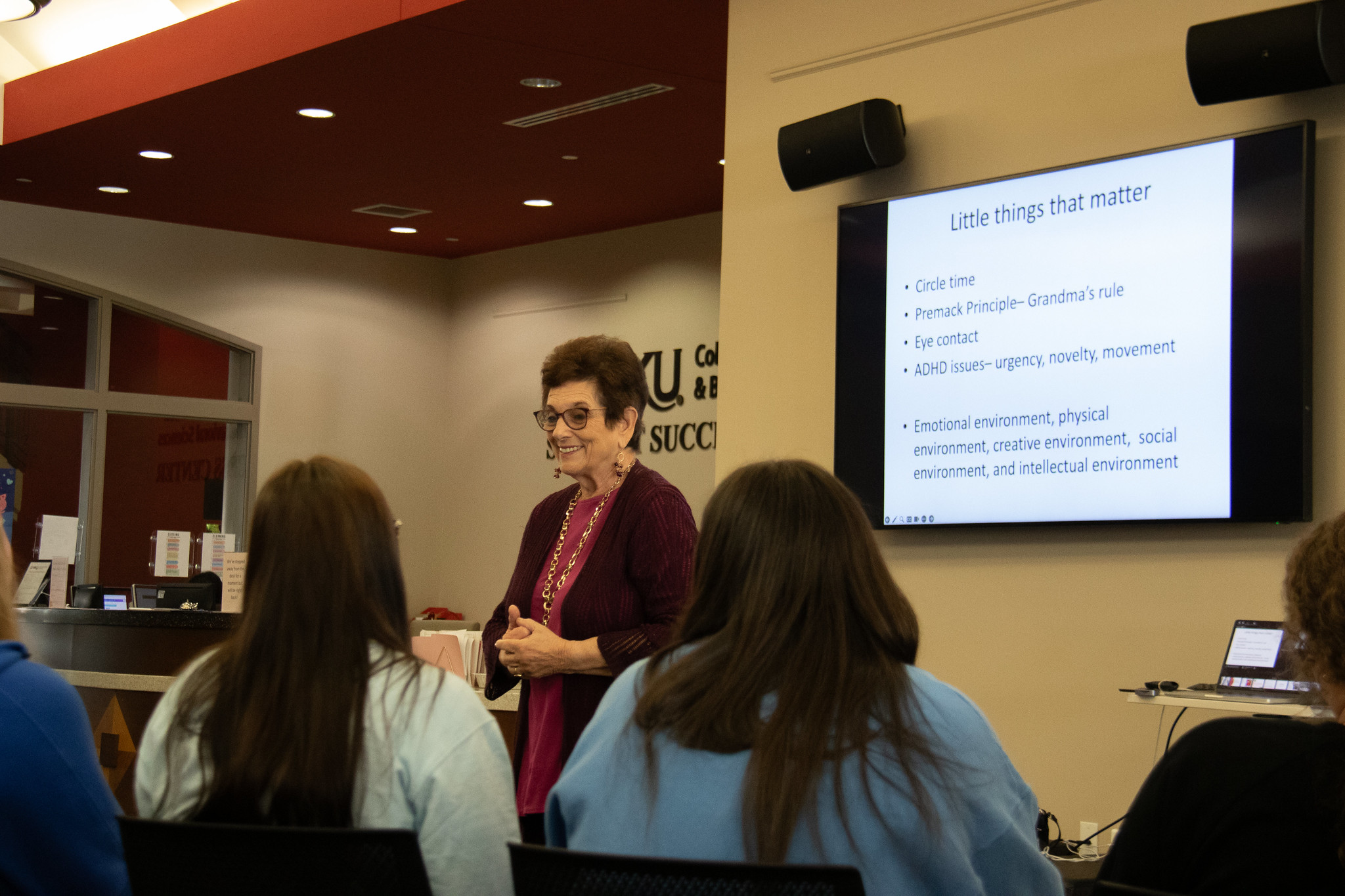 image of woman speaking in front of a group with a television behind her showing her slides