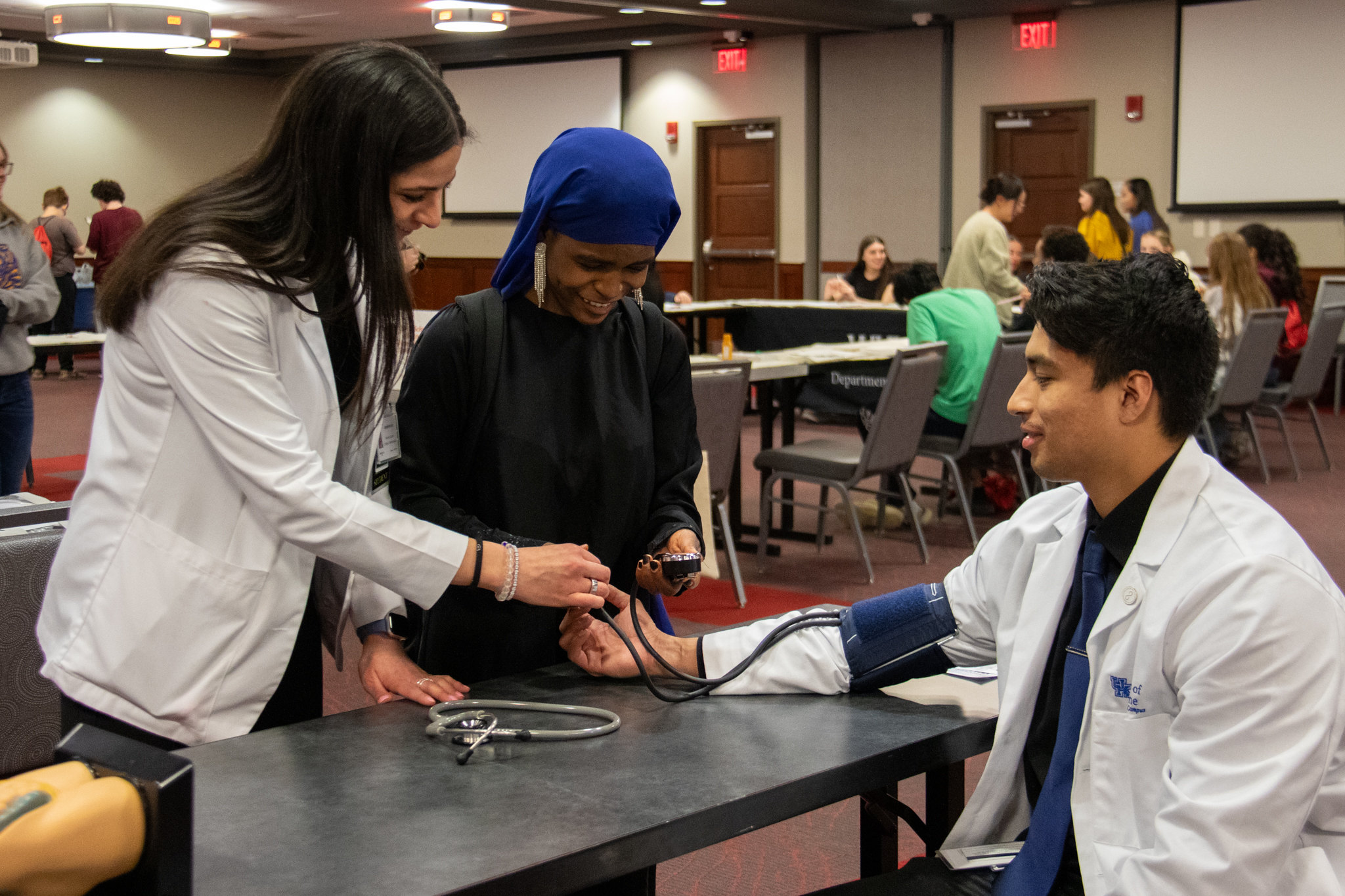Two college medical students in white jackets teach a middle school student how to take blood pressure readings