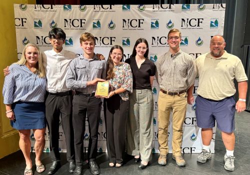 Group photo of Envirothon team and coaches with plaque standing in front of NCF banner.