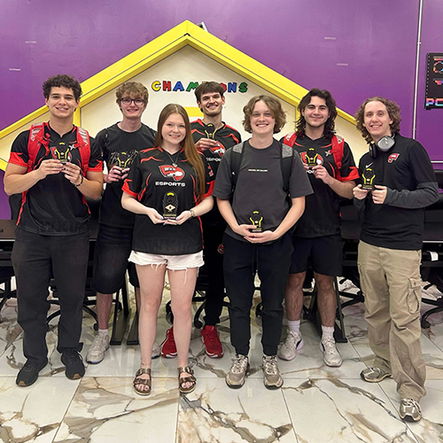 Seven WKU Varsity Esports students stand smiling, each holding a small trophy, in front of a yellow house-shaped
