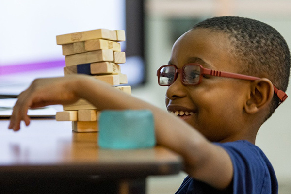 A smiling young boy wearing red glasses reaches toward a wooden block tower on a table during a Prime Time activity.