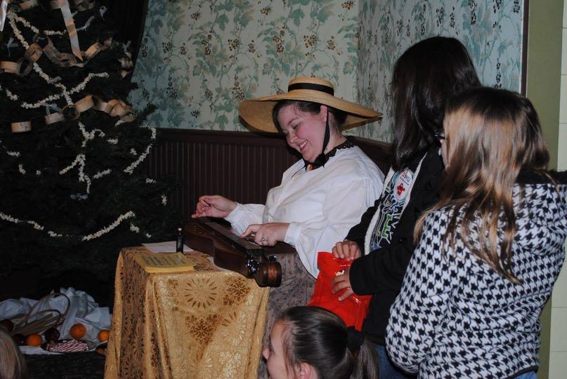 Woman in pioneer clothing, smiling and playing a dulcimer with children nearby