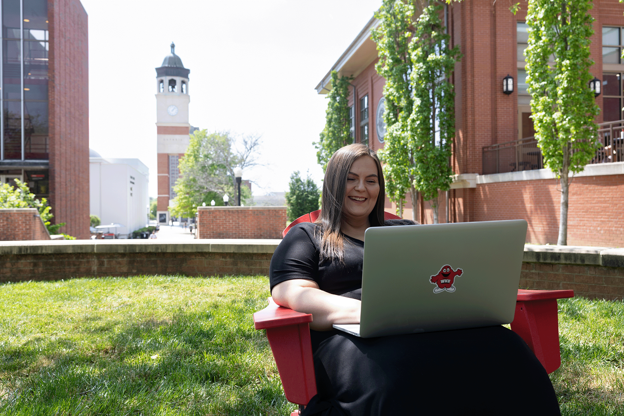 WKU student sits in a red chair with a laptop on a sunny day, near Guthrie Bell Tower on the WKU campus.