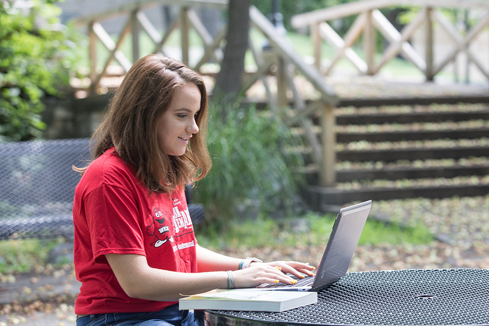 WKU student sits in a red chair with a laptop on a sunny day, near Guthrie Bell Tower on the WKU campus.