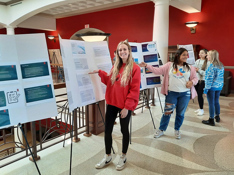 A psychology student points to her research poster while presenting findings to attendees at an academic conference.