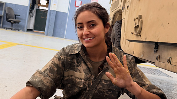 Cadet Harlow smiling and waving a grease-stained hand while sitting beside a military vehicle in a garage bay.