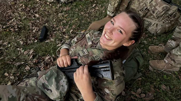 Cadet Reagan Abbott smiling while resting in the grass against field packs, holding a training rifle during an outdoor exercise.